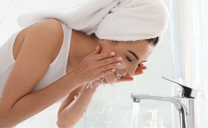 Woman with towel wrapped around her head washing her face with fresh water from a modern bathroom faucet.